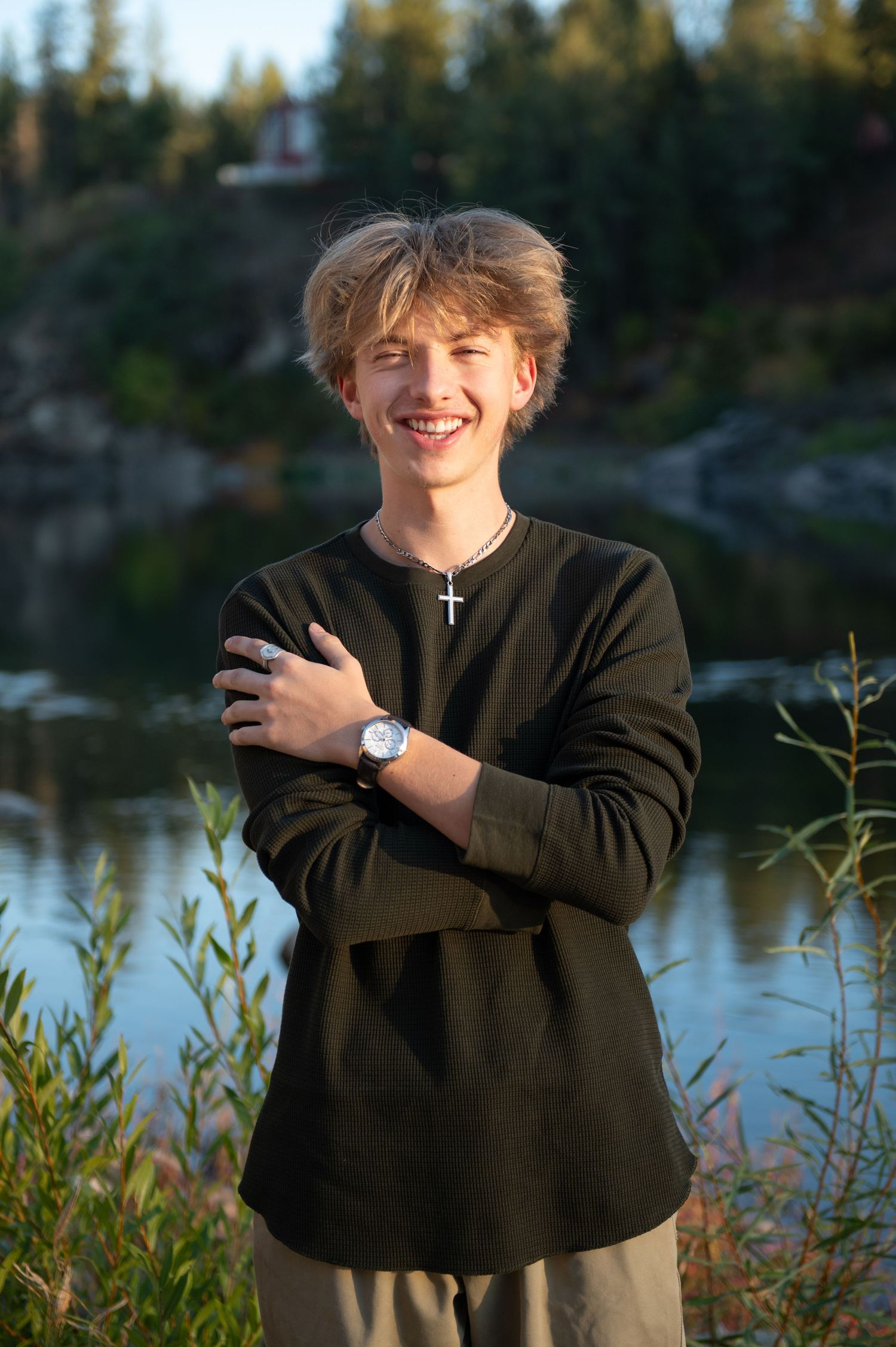 Smiling teenage boy with blonde hair wearing a dark sweatshirt and cross necklace, posing by a river at sunset.