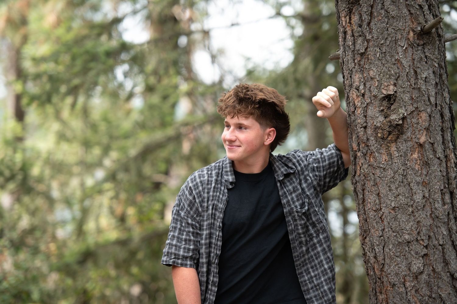 Teenage boy leans against pine tree smiling in forest setting.
