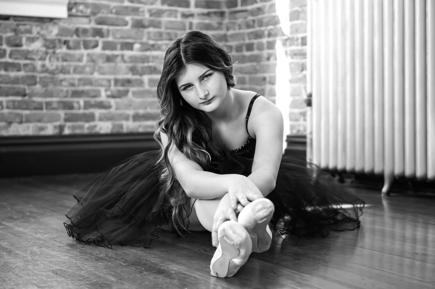Young ballerina in black tutu sitting on studio floor in black white photo.
