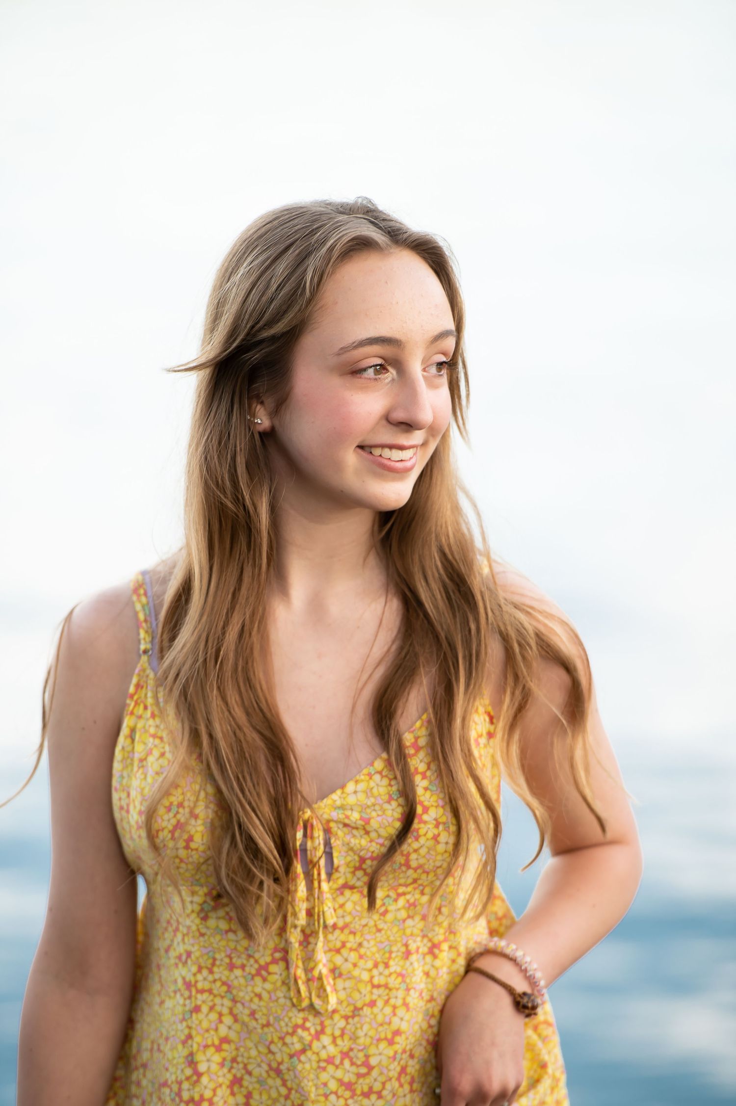 Smiling teenage girl in yellow floral spaghetti strap dress with long wavy hair outdoors near water.