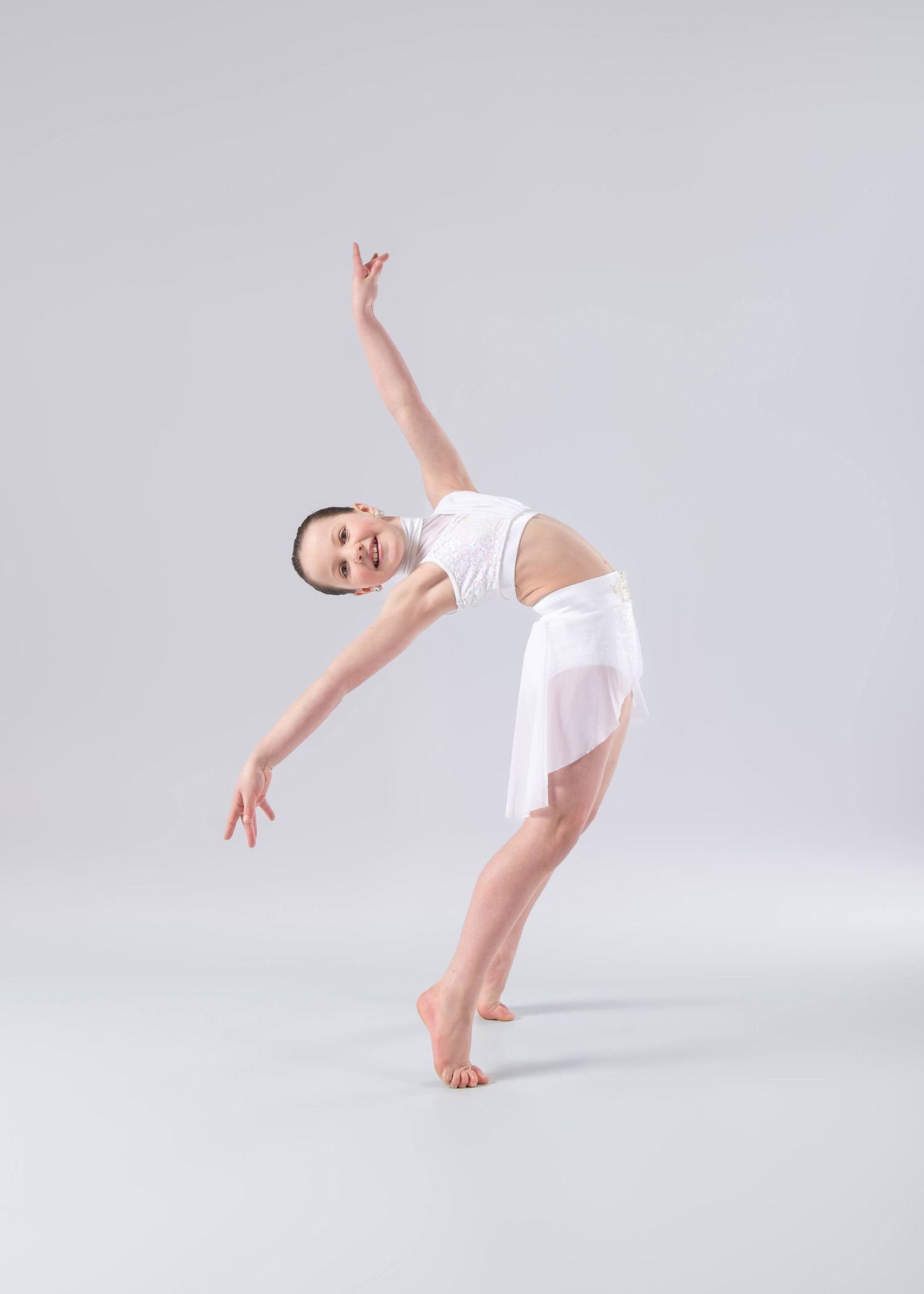 A young ballet dancer in white costume performs a graceful backbend with arms extended against a light gray background.