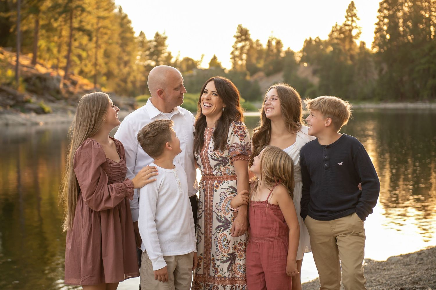 Happy family of six smiling together by a sunlit lake surrounded by tall pine trees at golden hour.