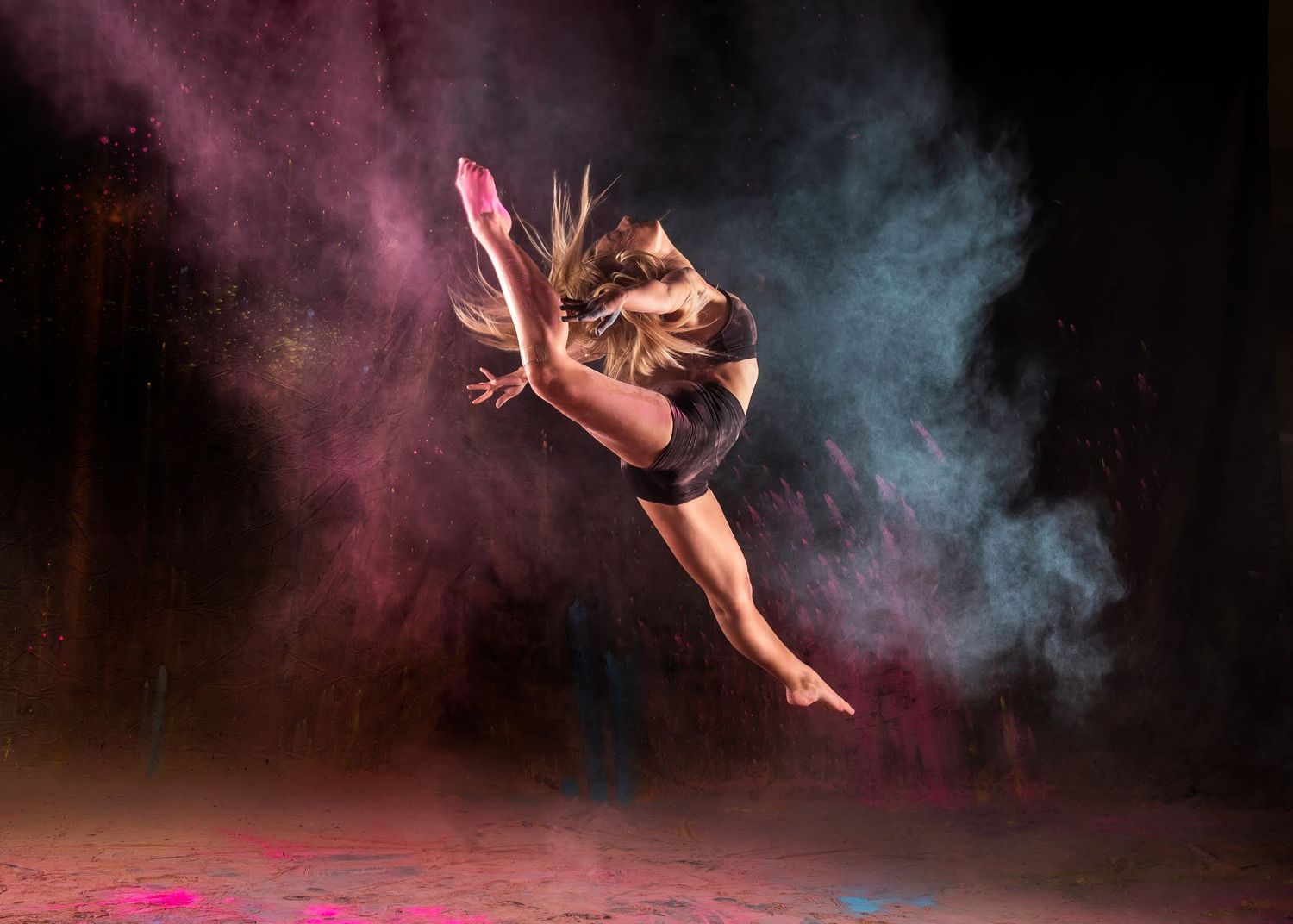 Female dancer leaping dramatically through colorful pink and blue powder cloud on dark stage.