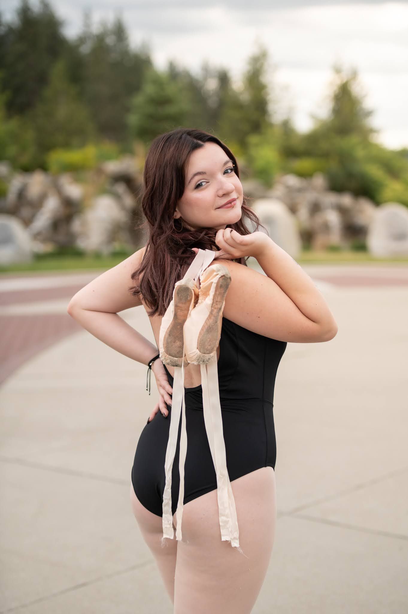 Young ballerina in black leotard holding pointe shoes over shoulder outdoors.