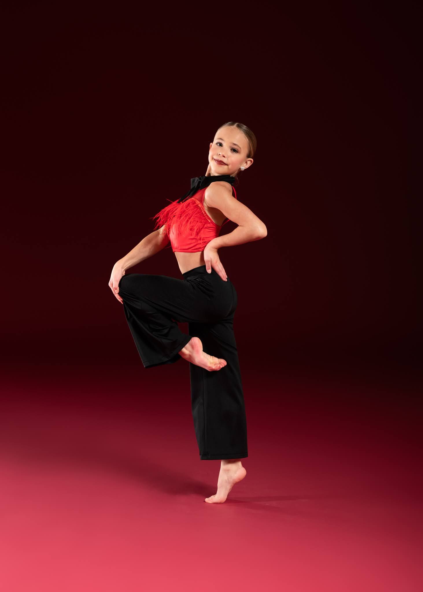 A young female dancer balances on one foot in a red crop top and black wide-leg pants on a red backdrop.