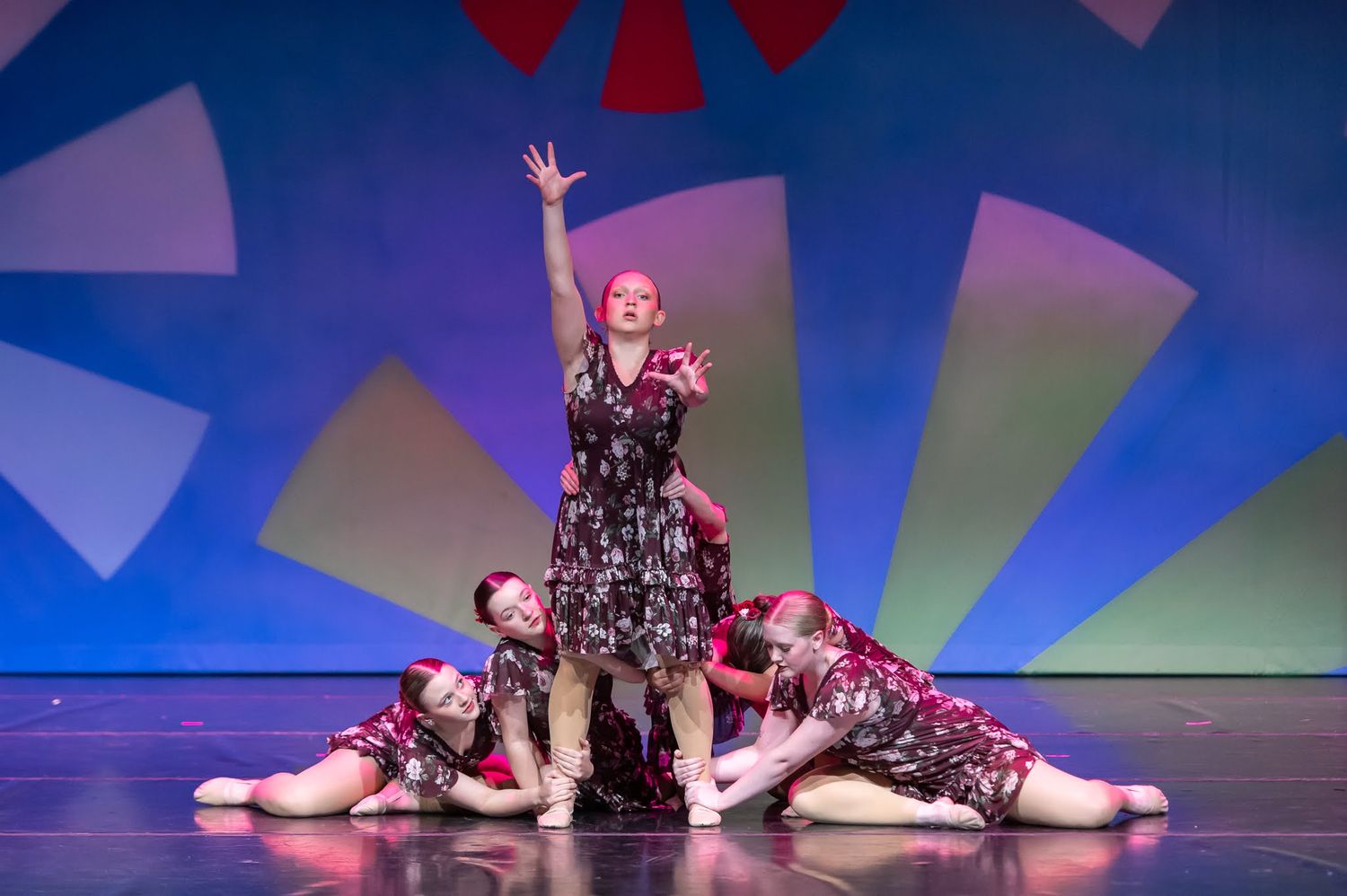 Group of dancers in matching floral costumes perform on stage with colorful geometric backdrop, one dancer raised above.