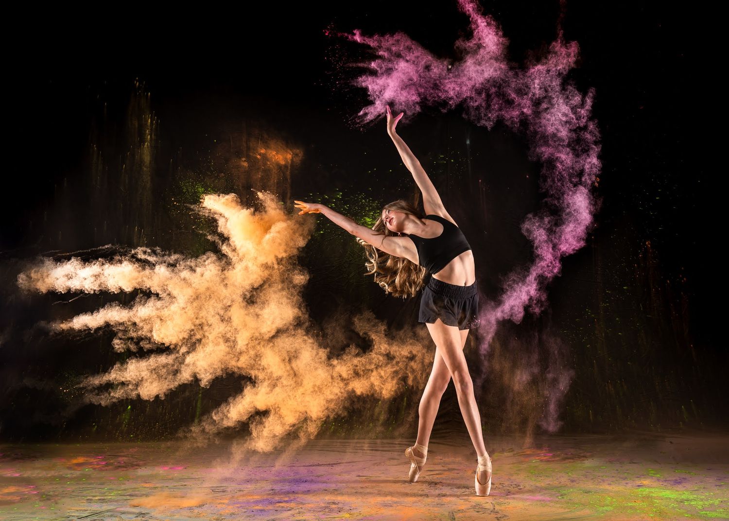 Ballerina dancing en pointe surrounded by swirling orange and purple powder explosion on dark stage.
