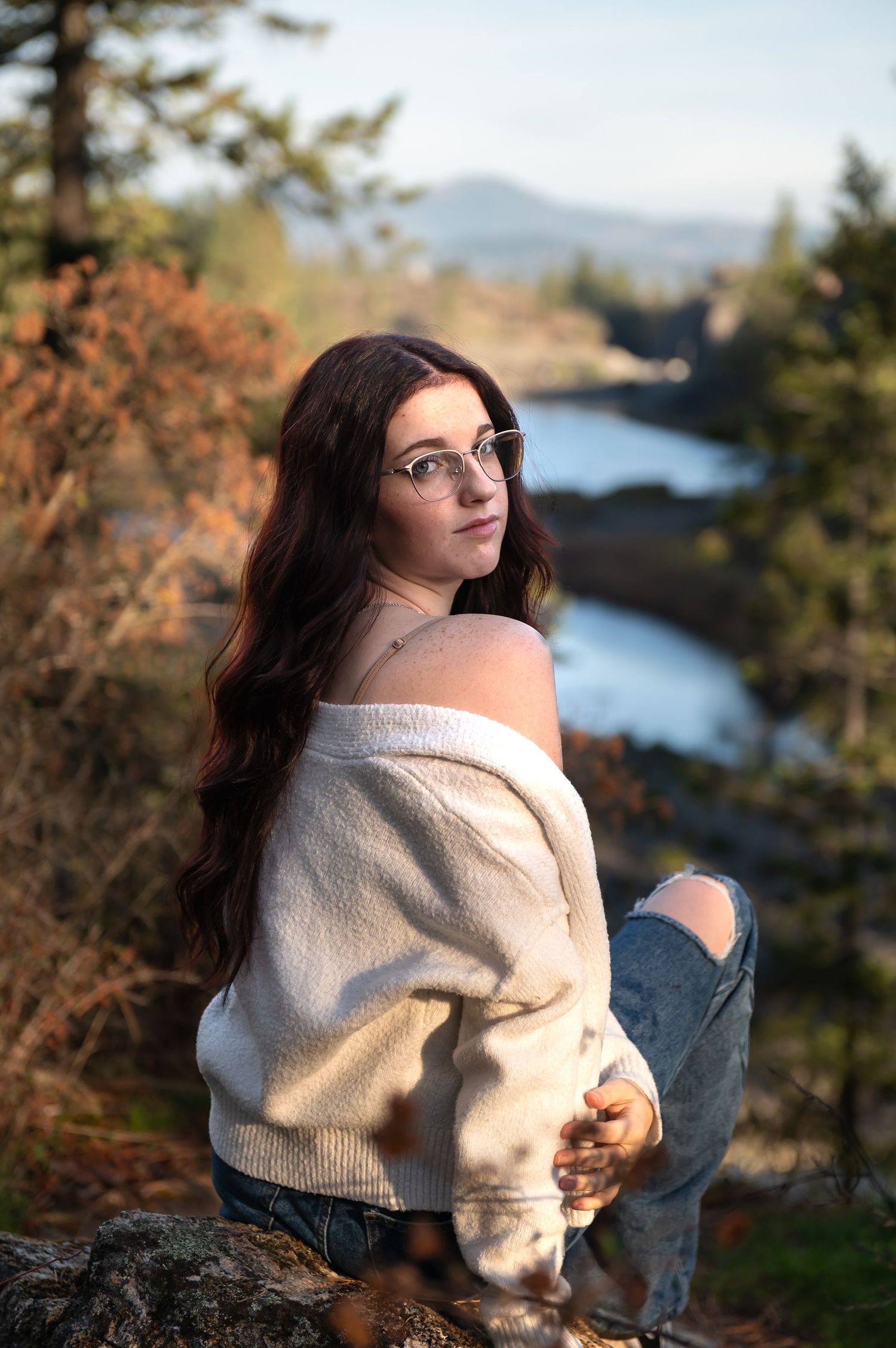 Brunette teen in off-shoulder sweater and glasses poses on rocks with scenic river view behind her.
