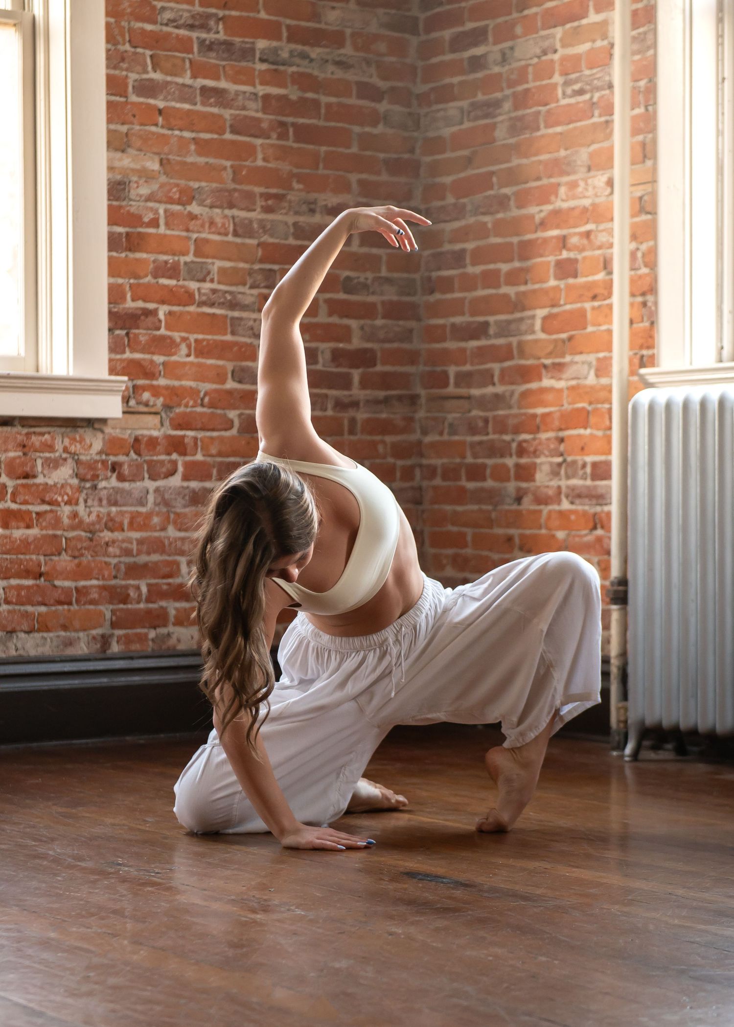 A dancer in white performs a graceful floor pose against a rustic brick wall in a sunlit studio.