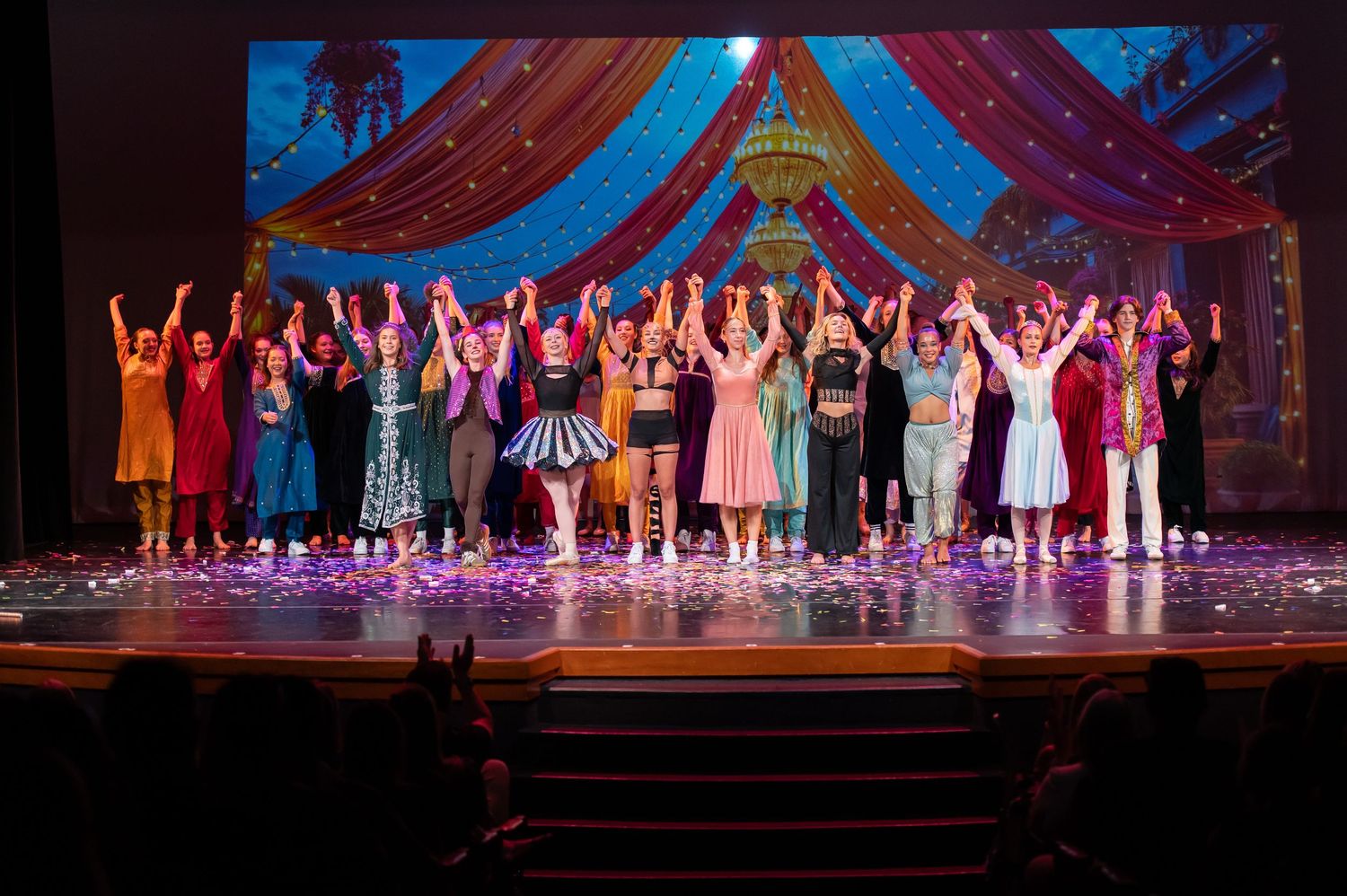 Large ensemble cast takes a final bow on stage with colorful backdrop and confetti.