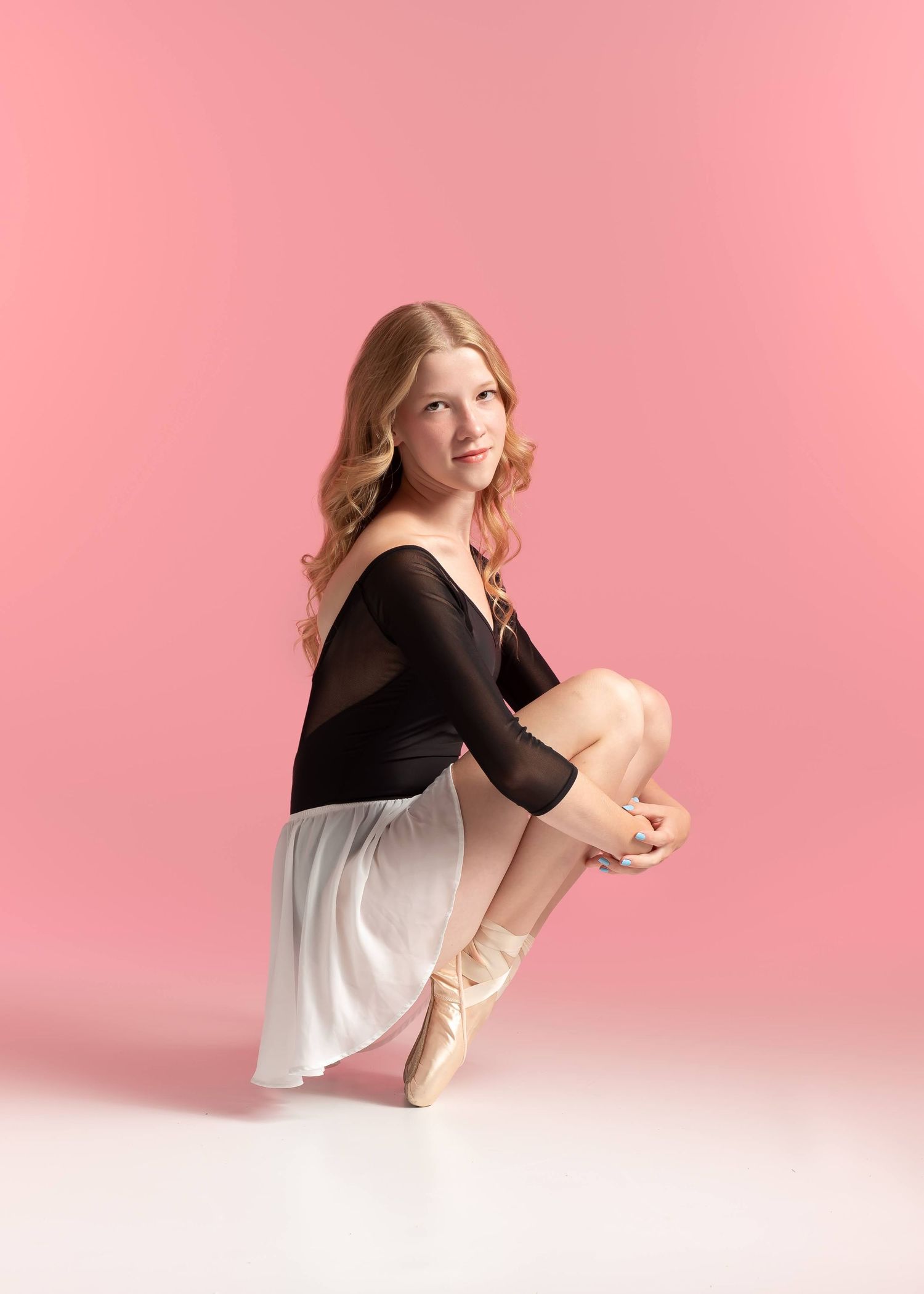 A young ballet dancer with red hair in a black leotard and white skirt sits gracefully on a pink background.