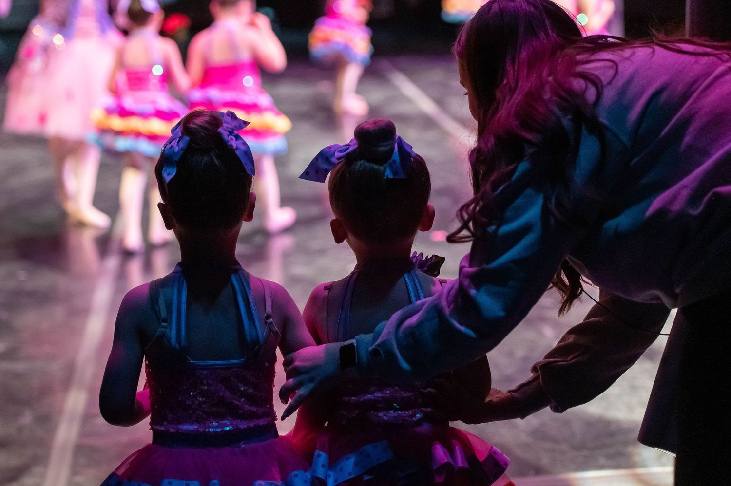 Adult helper adjusts costumes of two young ballerinas in pink tutus backstage under purple light.