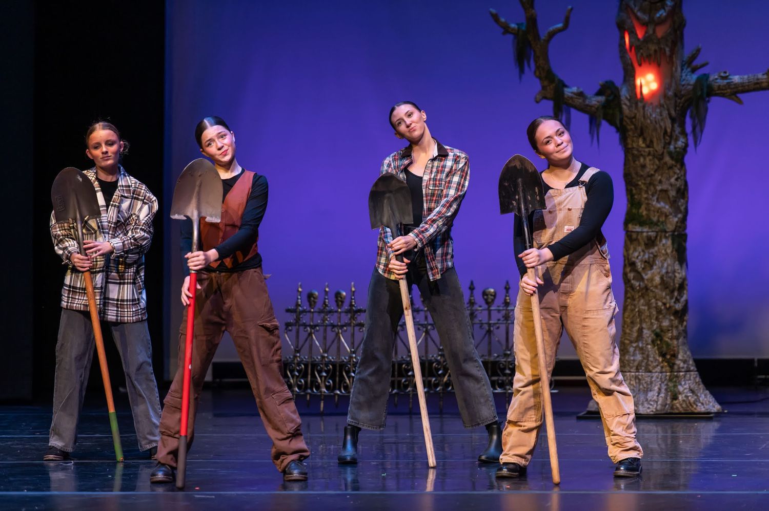 Four performers in costume holding shovels pose on stage with spooky tree and graveyard set design.