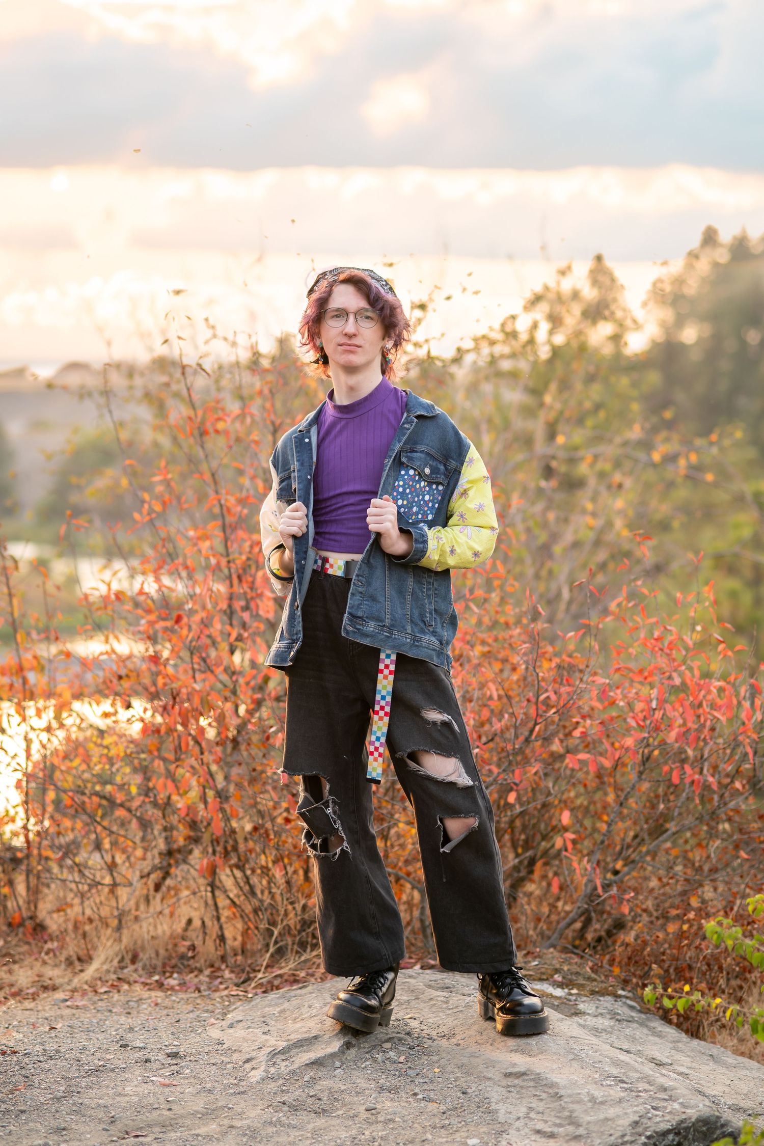 Young person in purple shirt, denim jacket, and ripped black pants standing on rock with autumn foliage backdrop.
