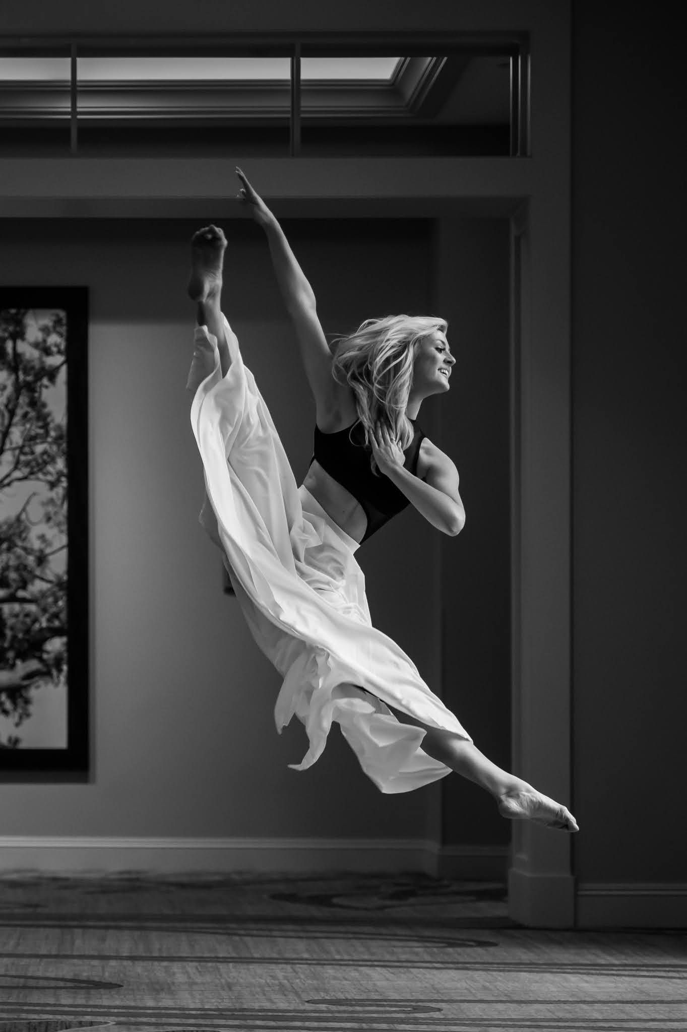 Black and white image of blonde dancer leaping mid-air with flowing white fabric in elegant split jump.