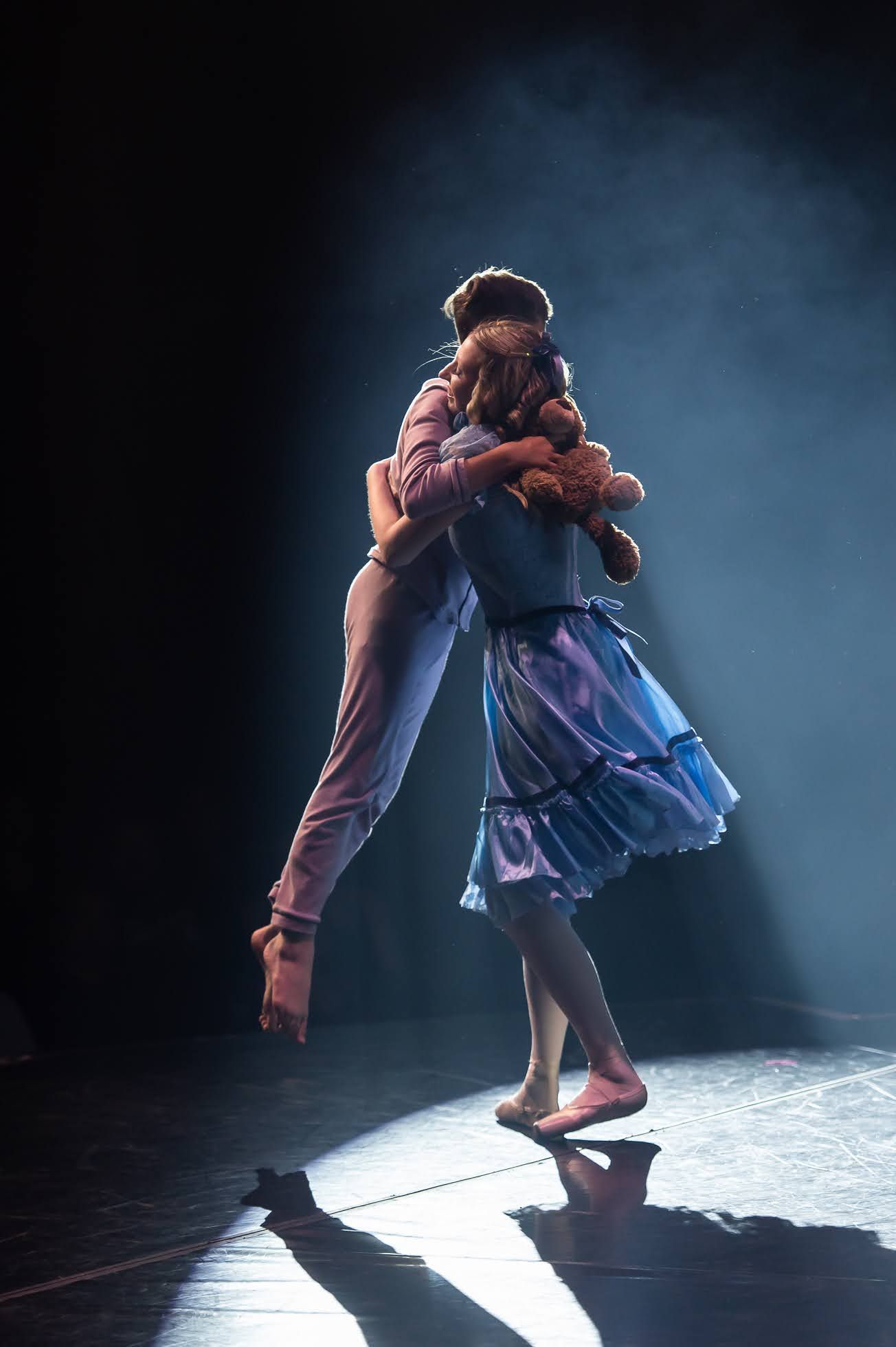 Two dancers share an emotional lift embrace under dramatic spotlight on a darkened stage.