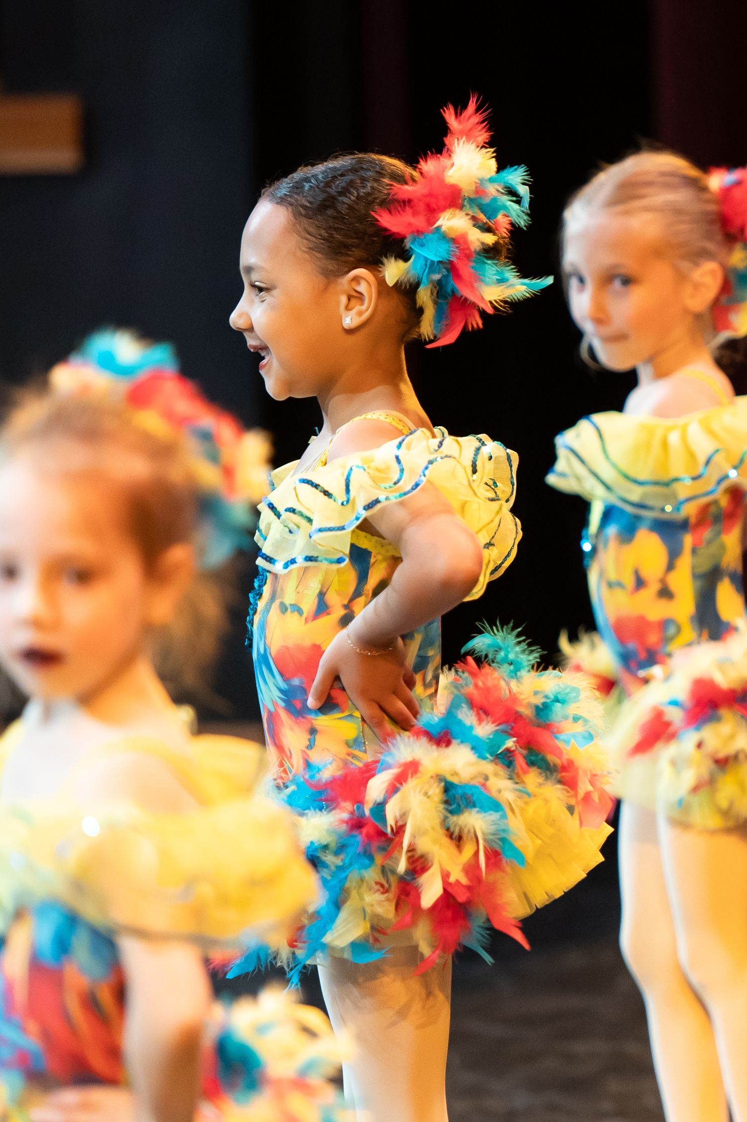 Young girls in colorful feathered yellow costumes perform in a dance recital on stage.
