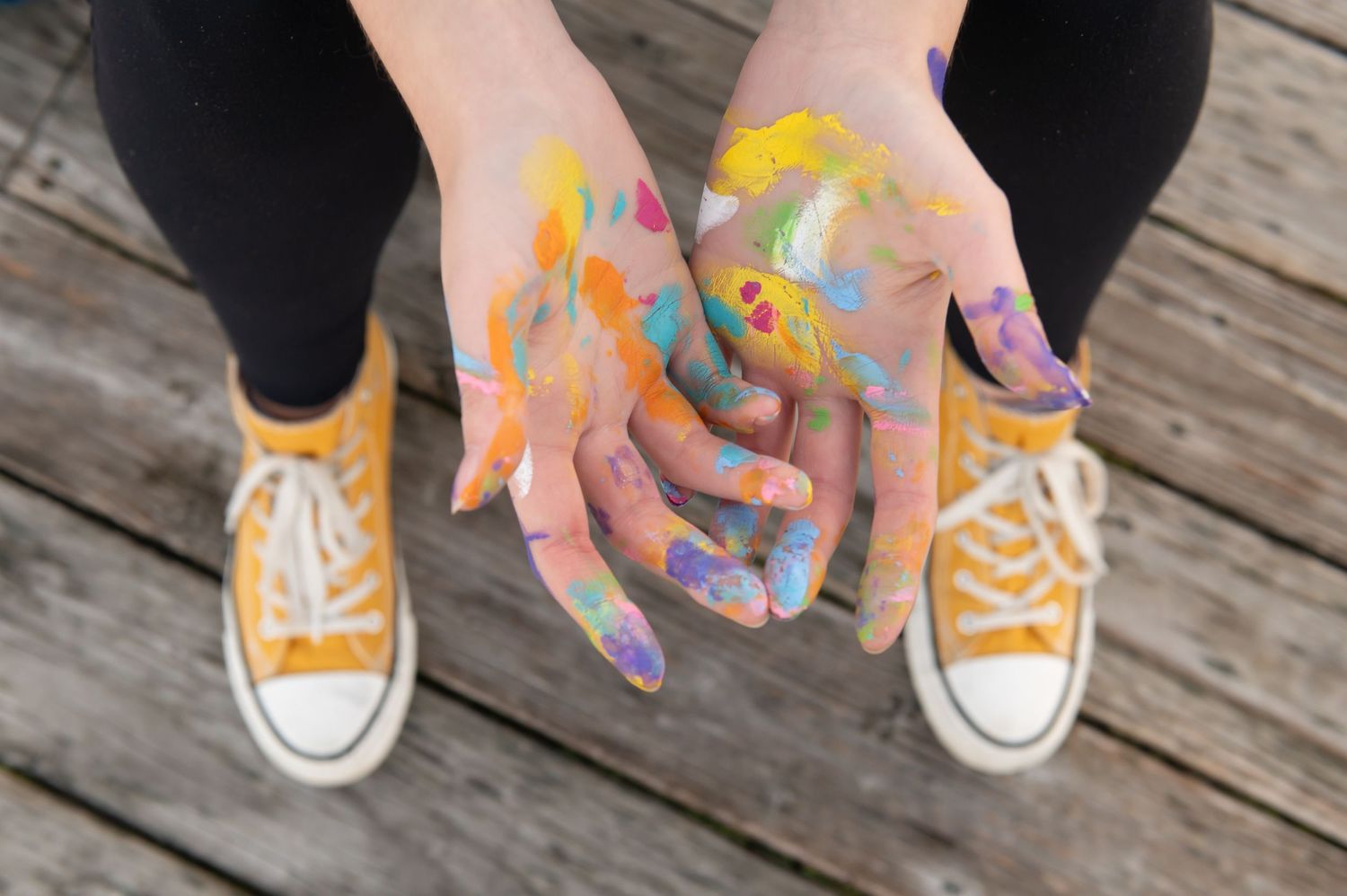 Close-up of hands covered in colorful paint held out over wooden dock with yellow sneakers visible.