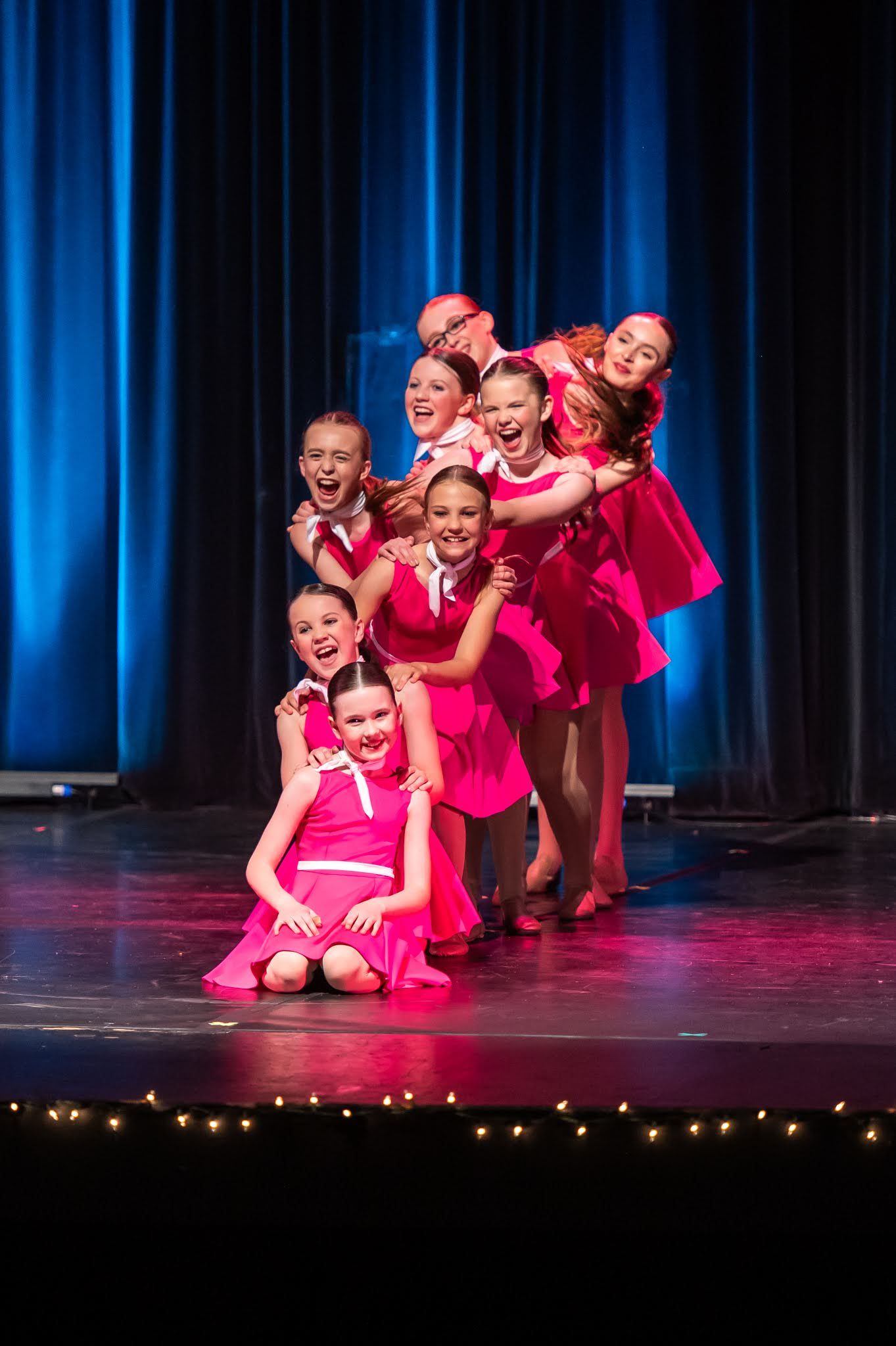 Young girls in pink dresses perform an energetic group dance routine on a theatrical stage.