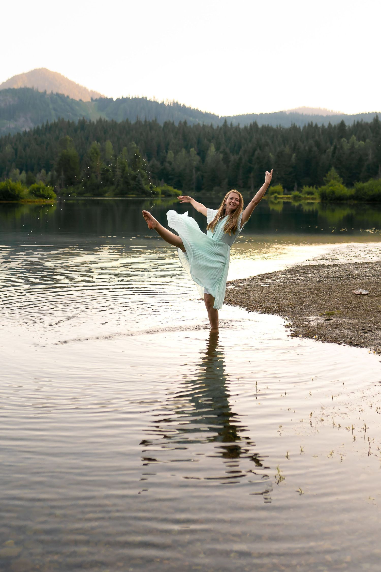 Blonde dancer in light blue dress performs arabesque in mountain lake at sunset.