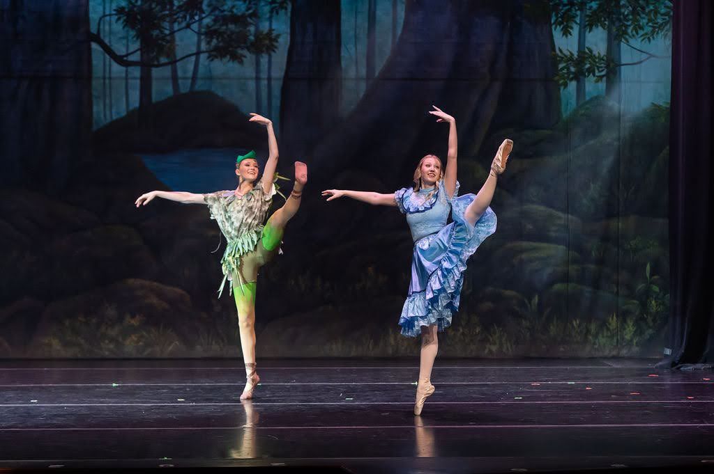 Two ballet dancers in Peter Pan costumes performing arabesques on stage with forest scenery.