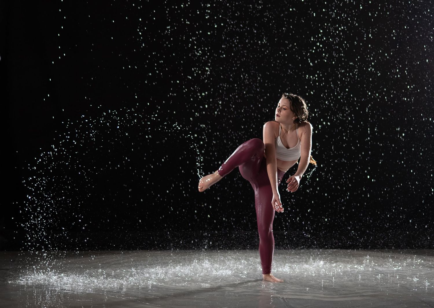Female dancer performing high kick in maroon leggings amid dramatic water droplets on dark studio background.