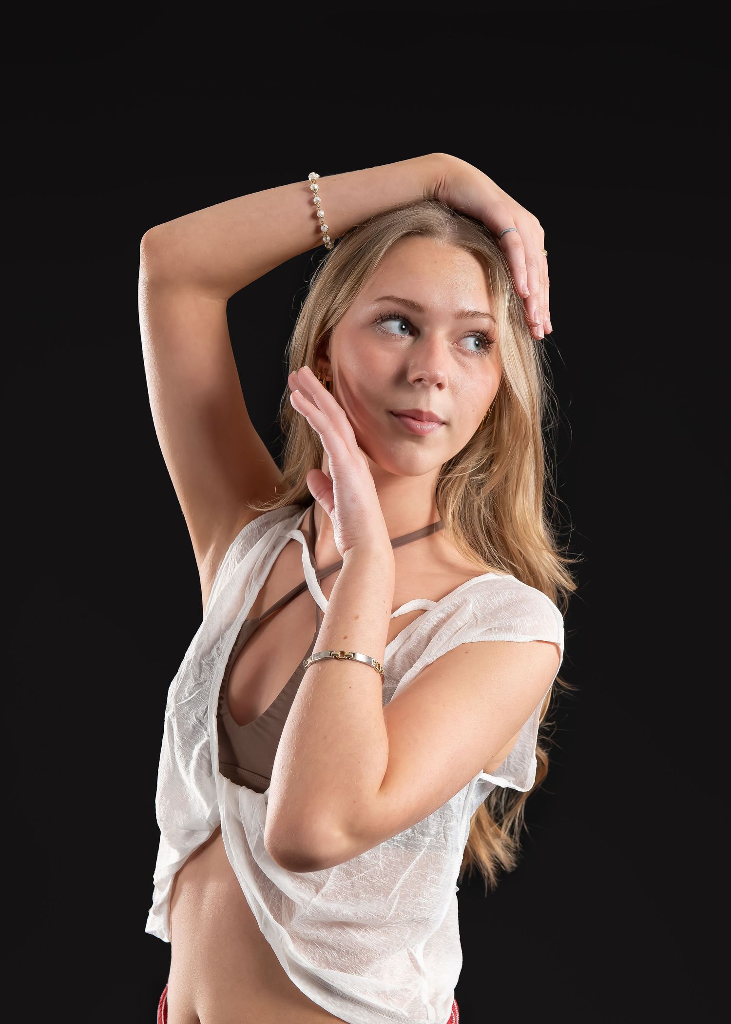 Young blonde female dancer poses gracefully with arms framing face on black background.