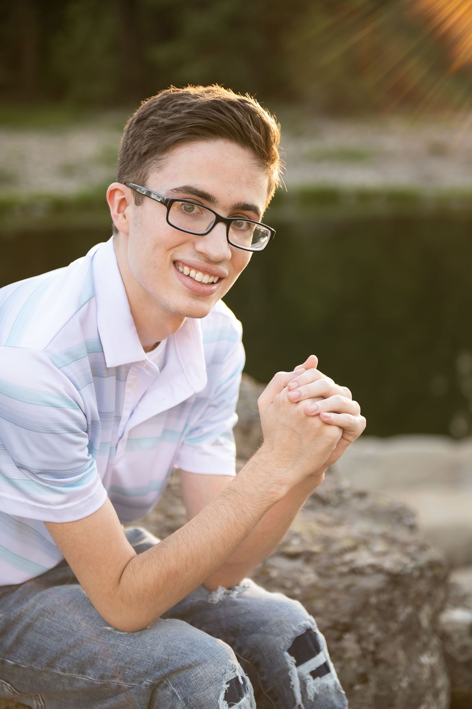 Smiling teen boy with glasses sits on rocks with clasped hands near a sunlit pond.