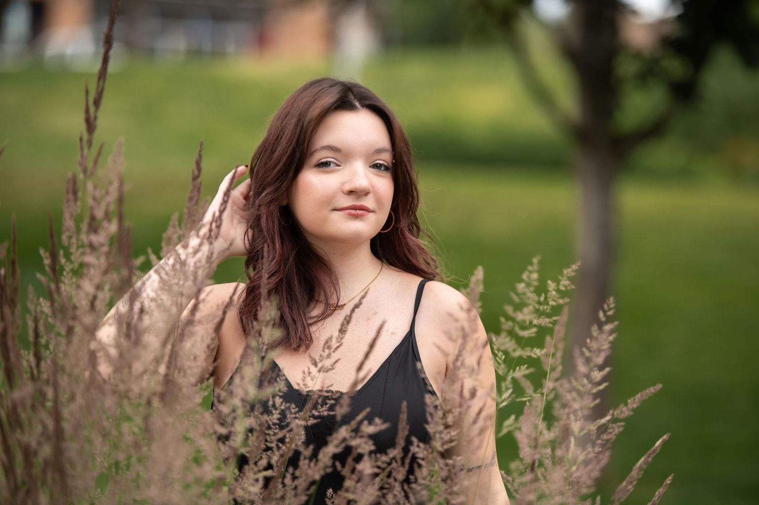 Young woman with auburn hair standing among tall grasses in a lush green outdoor setting.