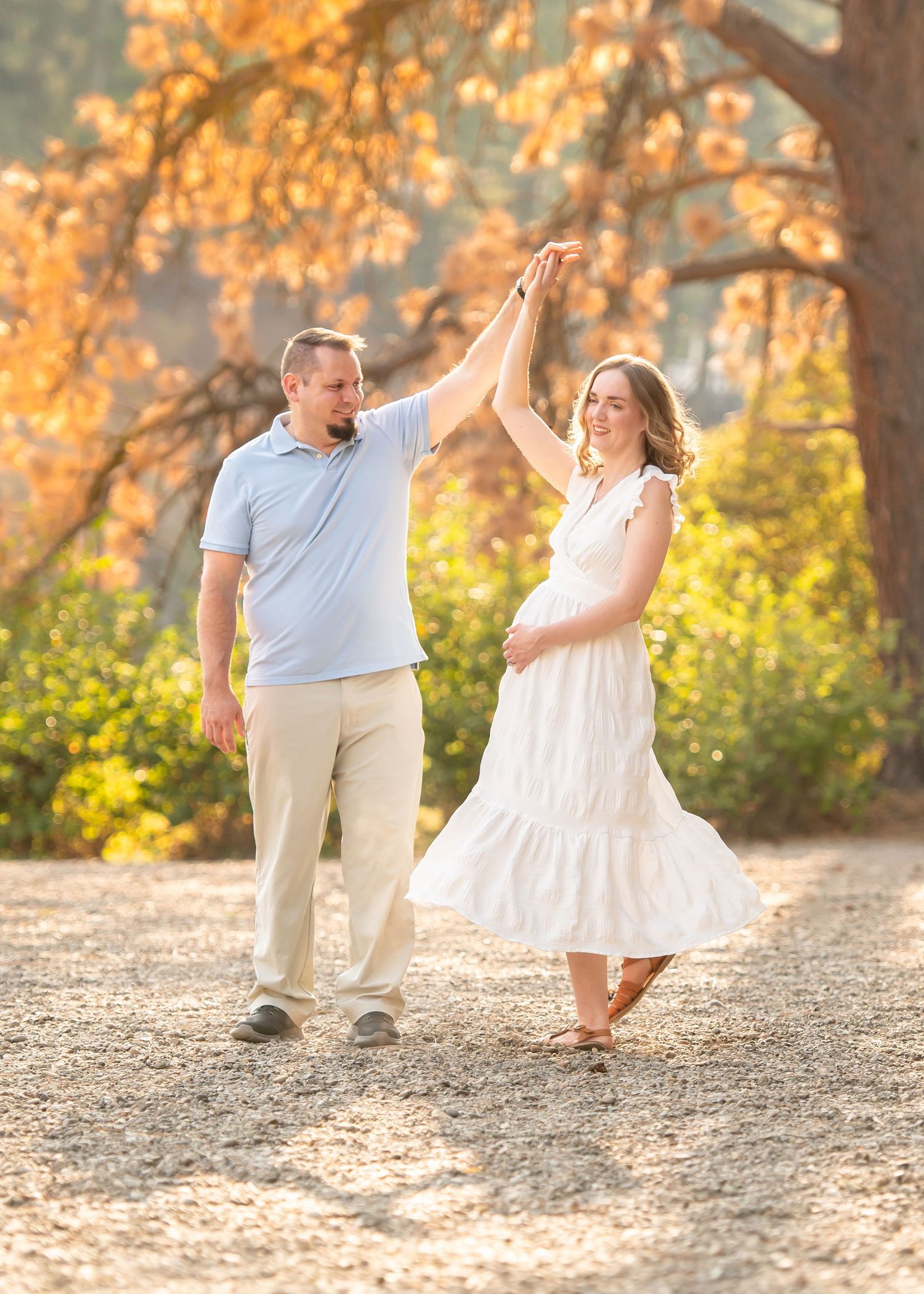 Happy couple dancing outdoors in golden autumn light, woman in white dress being twirled by man in light blue shirt.