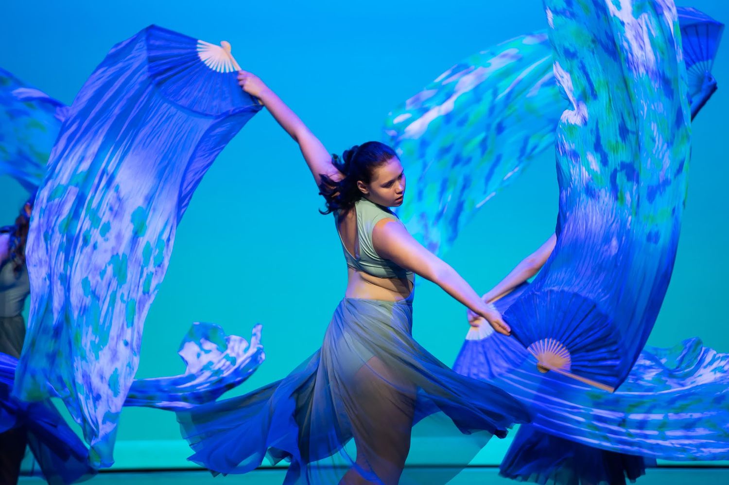 A dancer performs with large blue flowing fan veils under vibrant blue stage lighting.