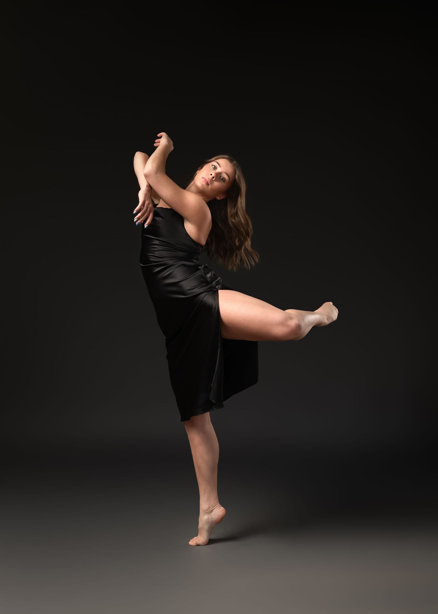 A female dancer in a black dress elegantly kicks her leg high in a dramatic contemporary dance pose against a dark background.