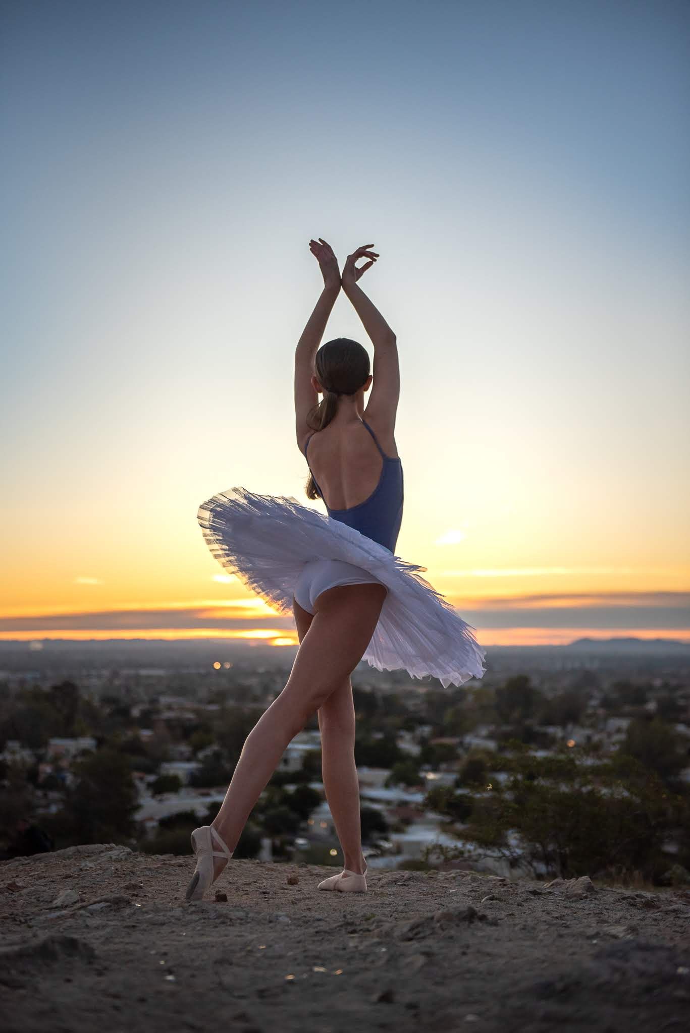 Ballerina in a white tutu and blue leotard poses on pointe at sunset with a city skyline backdrop.