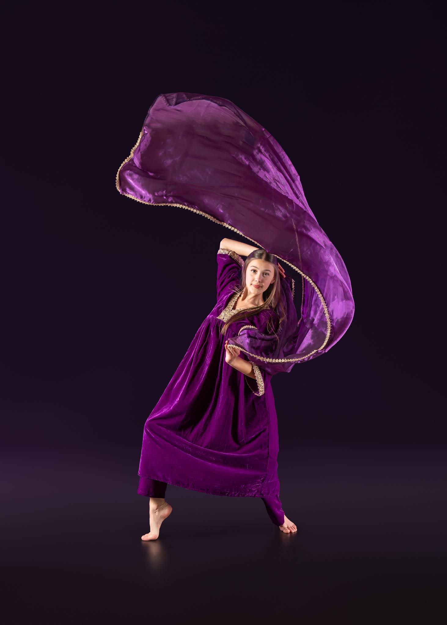Dancer in purple velvet gown tosses flowing violet fabric overhead on dark stage.