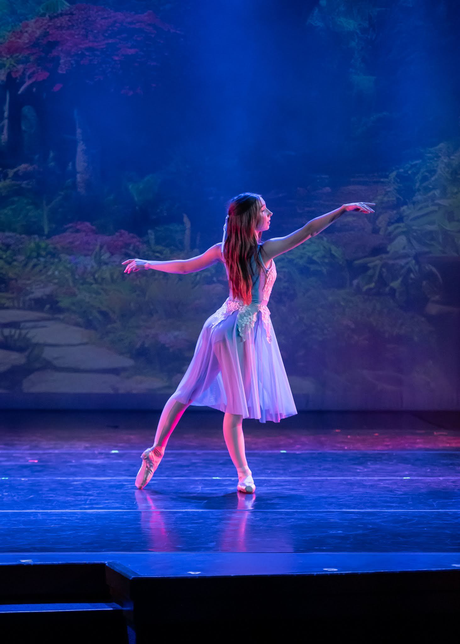 Ballerina in white tutu dress performs elegant en pointe pose under purple stage lighting.