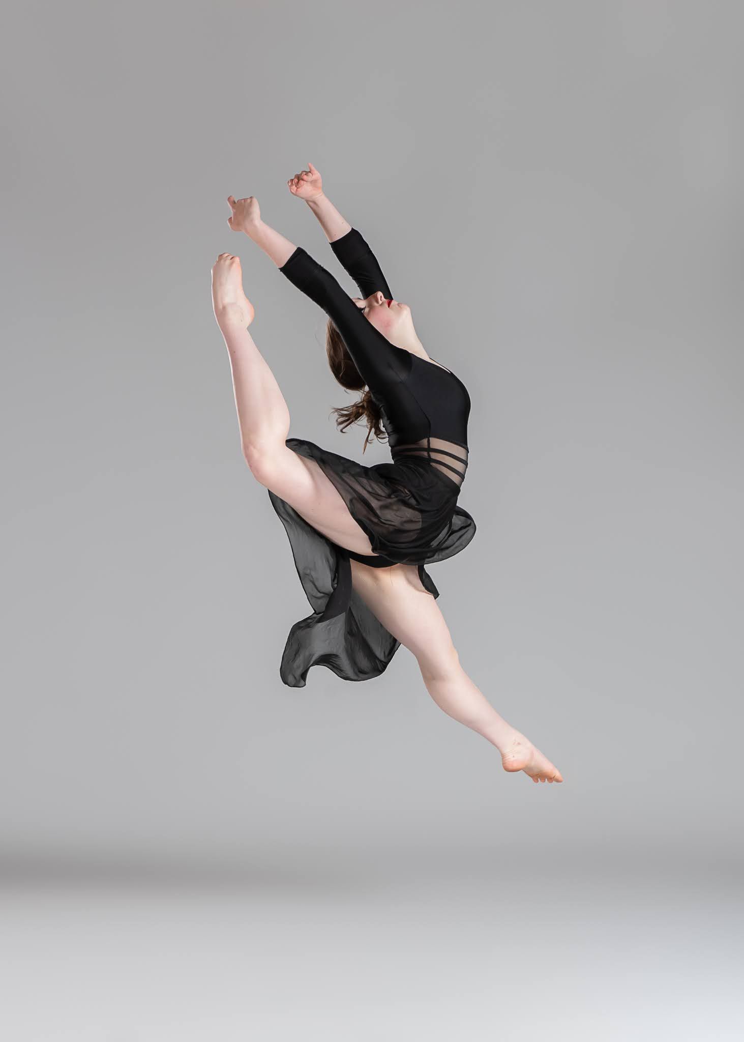 Dancer leaps gracefully in black costume with flowing skirt against gray studio background.