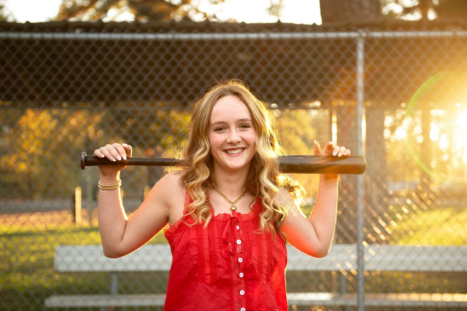 Smiling teenage girl in red dress holding a baseball bat across her shoulders on a sunny baseball field.