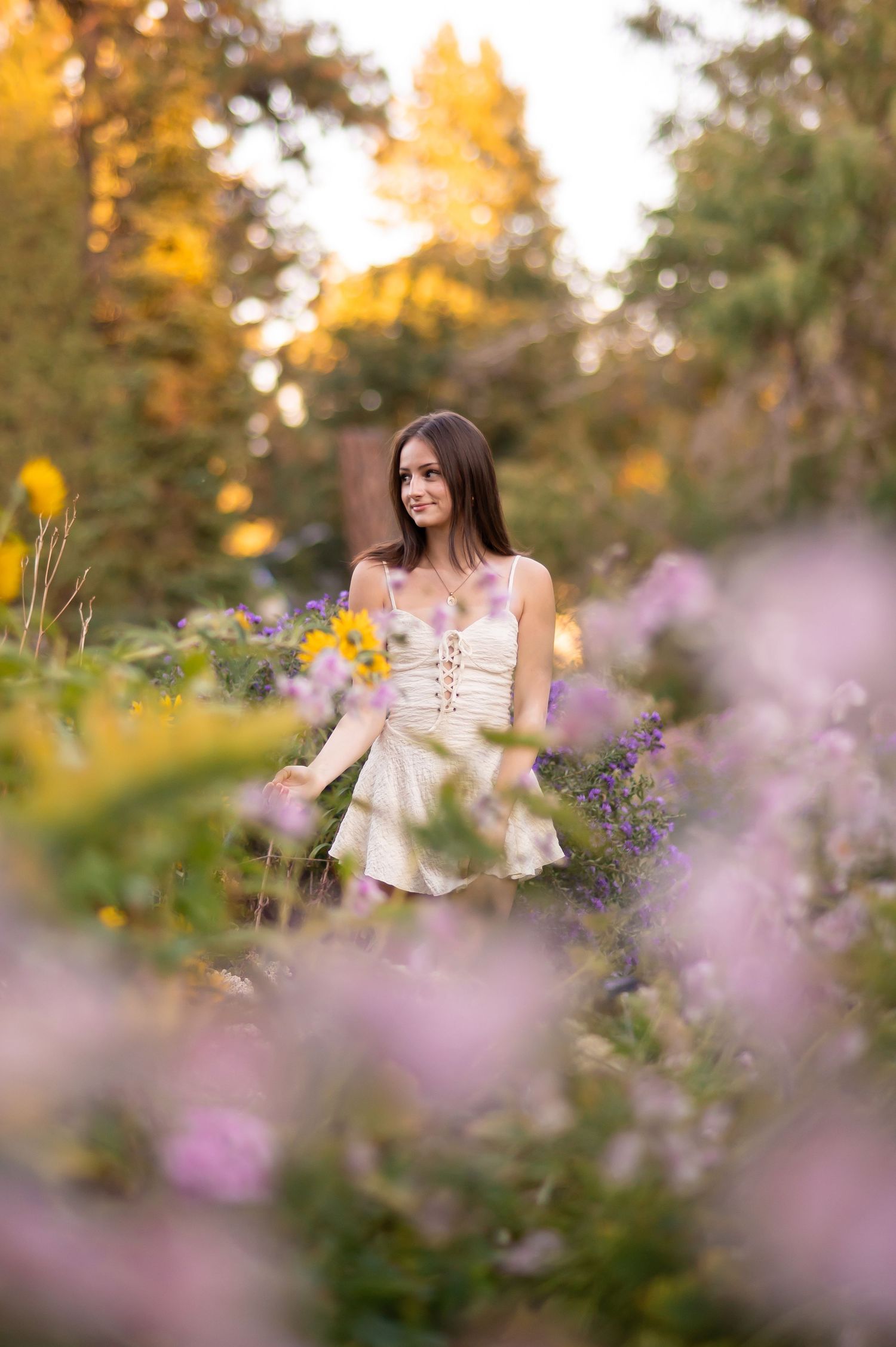 Young woman in a white dress standing among colorful garden flowers at sunset.