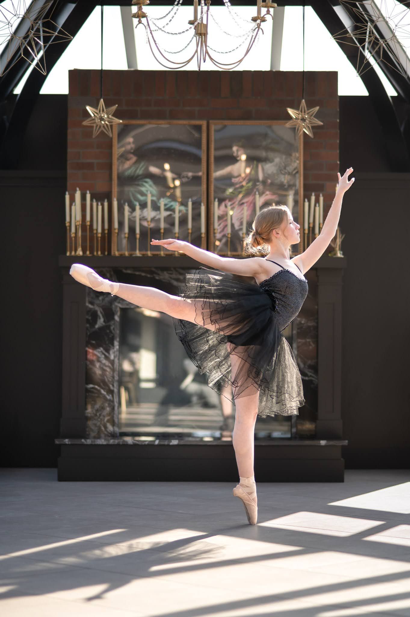 Ballerina in black tutu performing arabesque en pointe in elegant glass-roofed venue with candles.