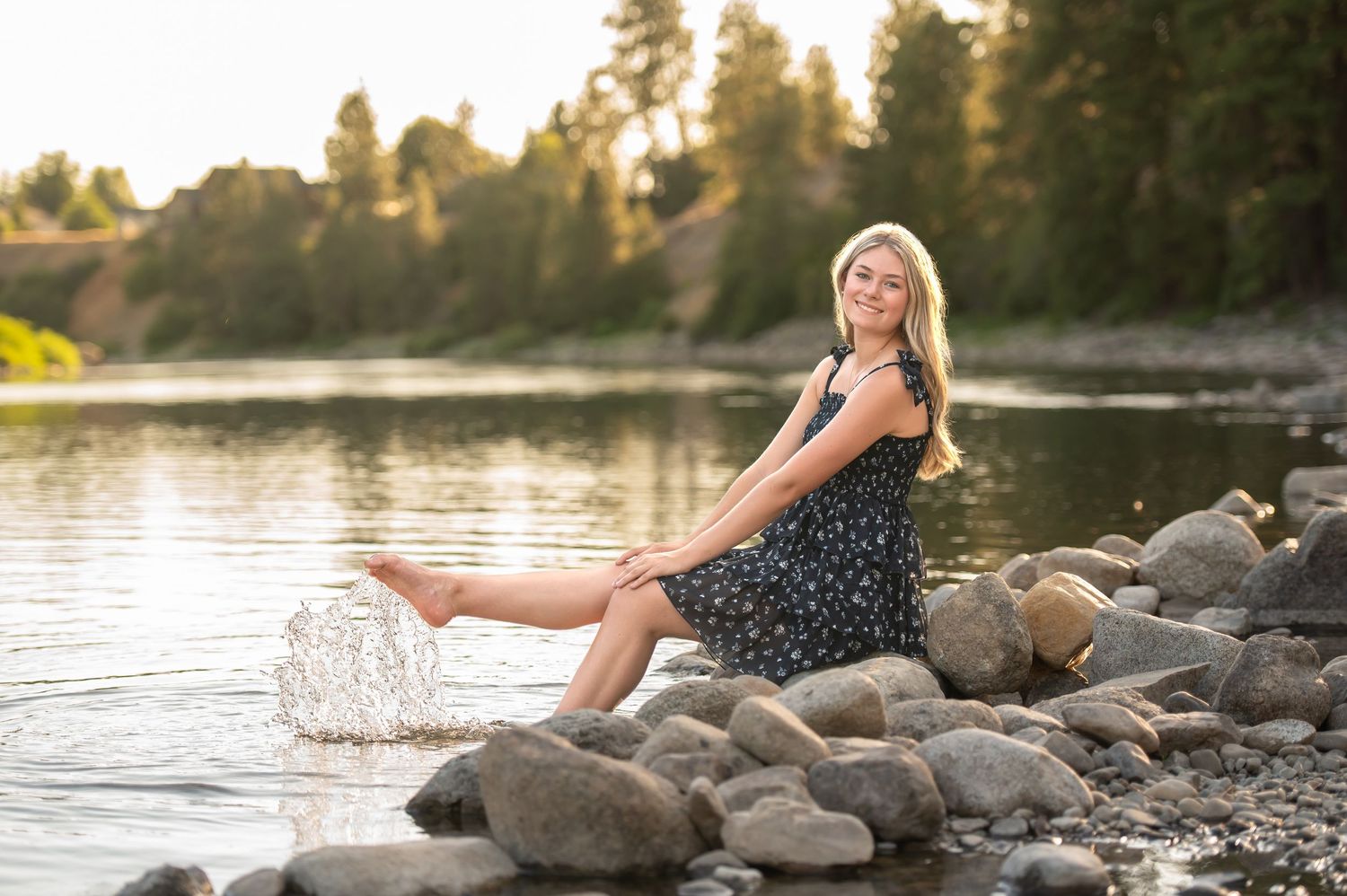 Blonde girl in floral dress sits on rocks splashing water at sunset.