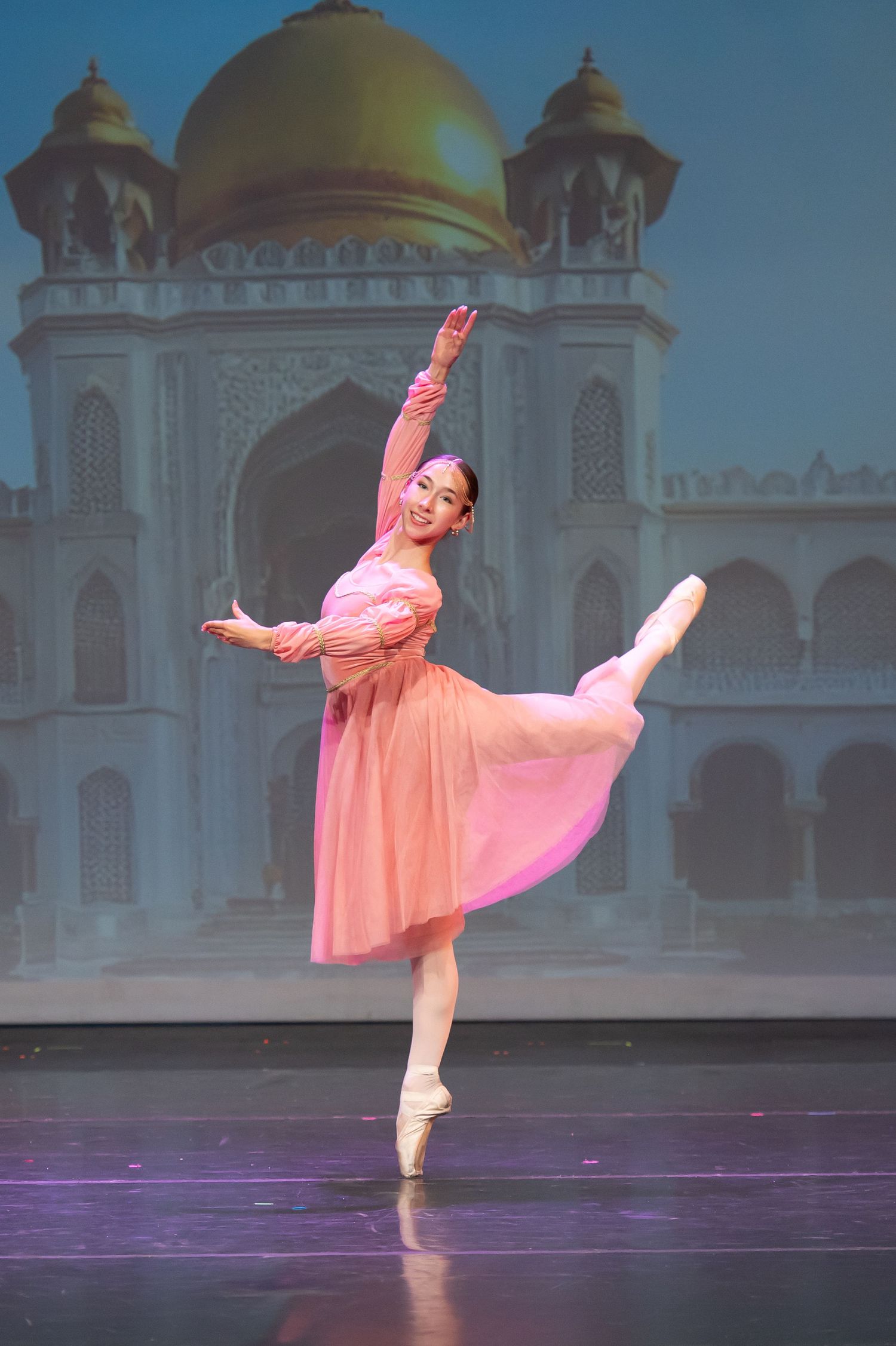 Ballet dancer in pink dress performs an arabesque on pointe before an Indian palace backdrop.