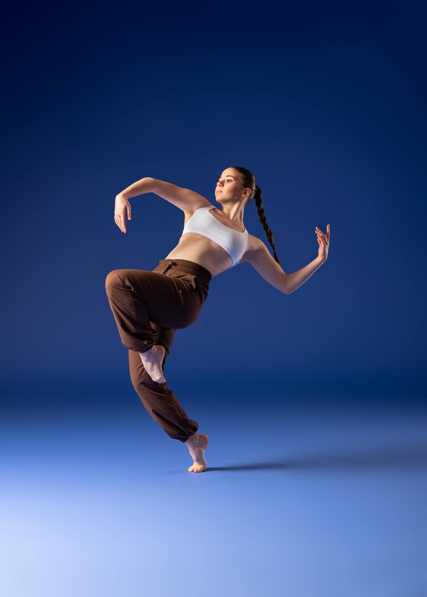 A contemporary dancer in white crop top and brown pants leaps dynamically against a deep blue studio background.