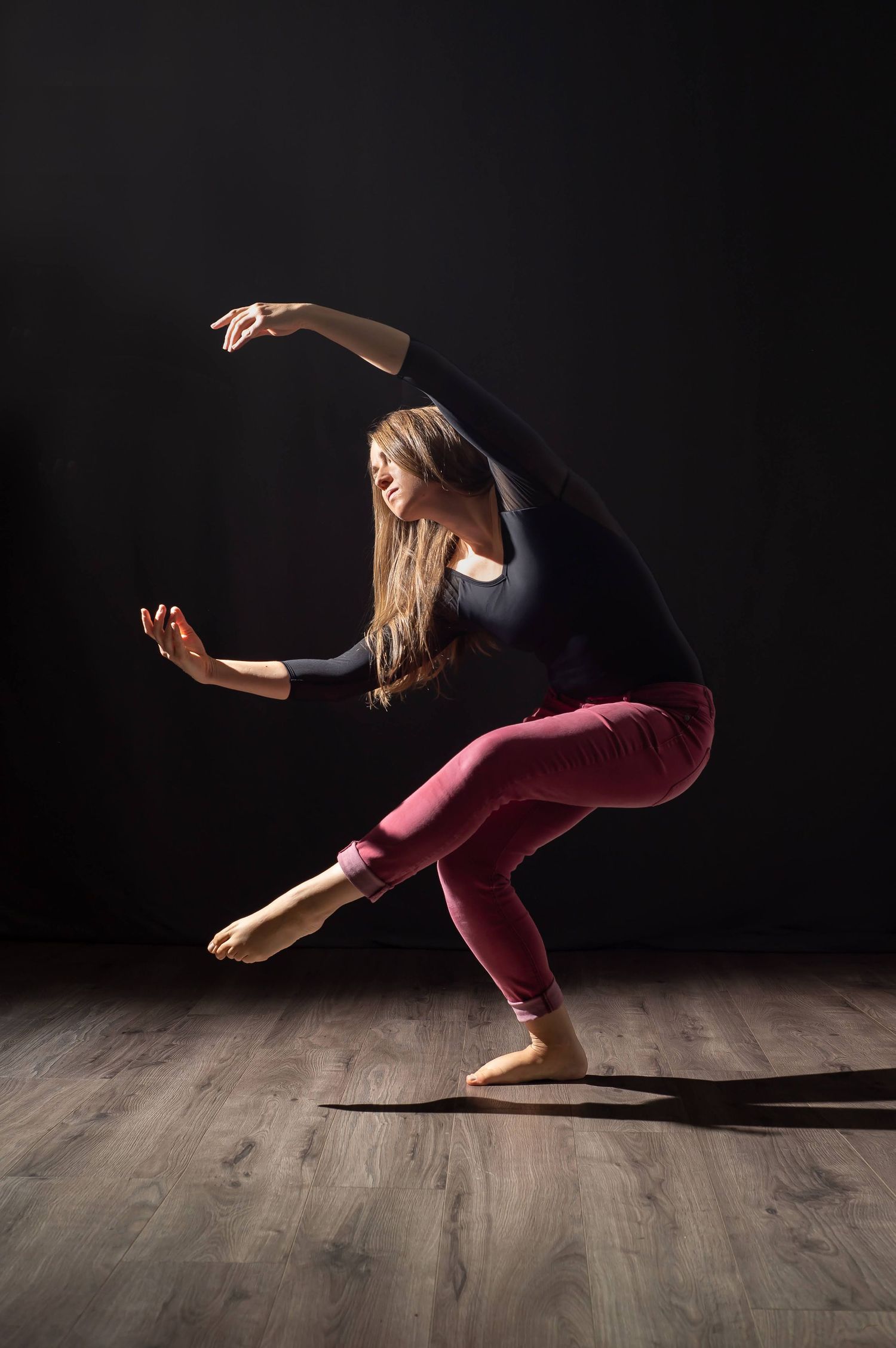 Charlotte Gardunia in a dramatic low squat pose with arms raised in a dark studio.