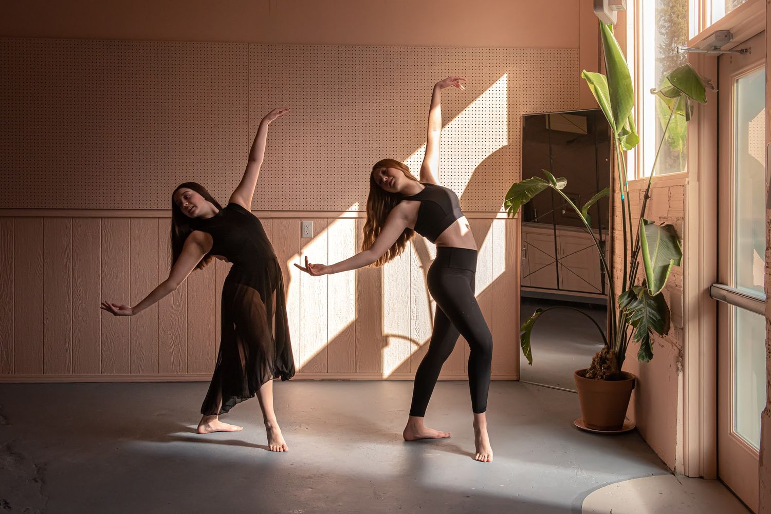 Two female dancers in black attire performing contemporary dance poses in sunlit studio with warm tones.