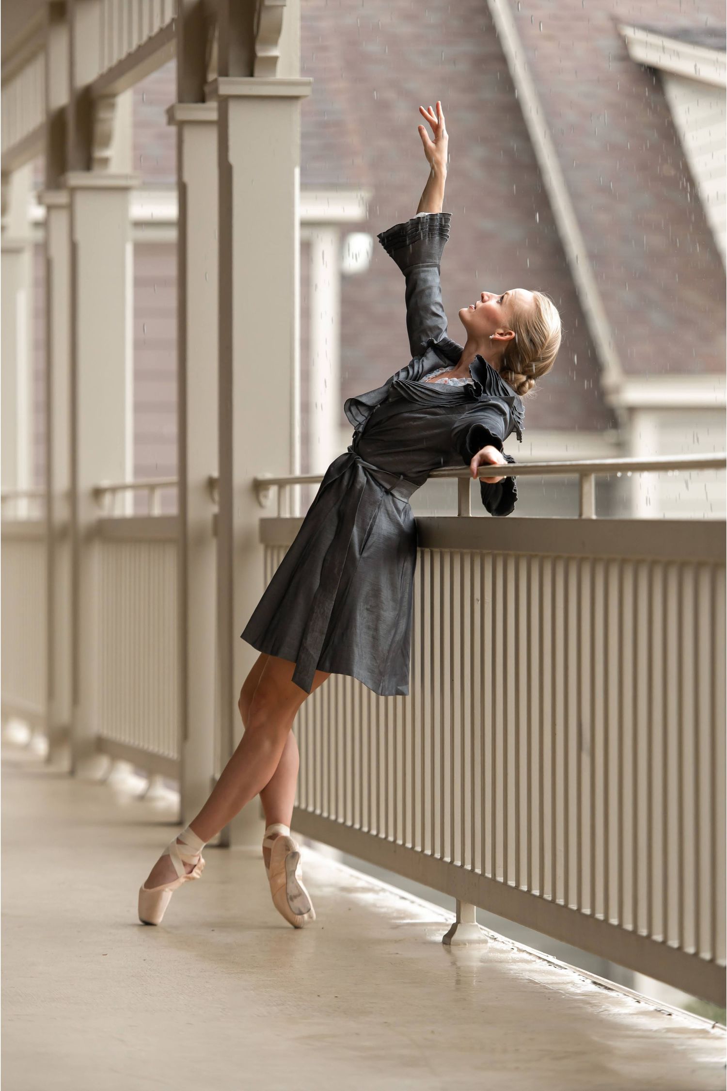 Ballerina in gray dress dancing en pointe on outdoor balcony railing with one arm raised gracefully watching rain fall.