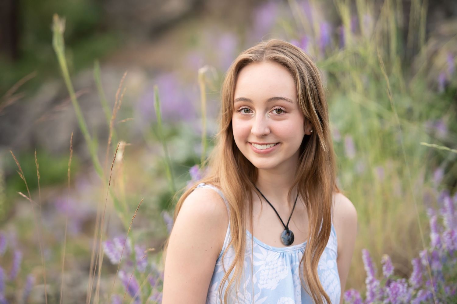 Smiling blonde teen girl sits among purple wildflowers in a serene outdoor portrait setting.