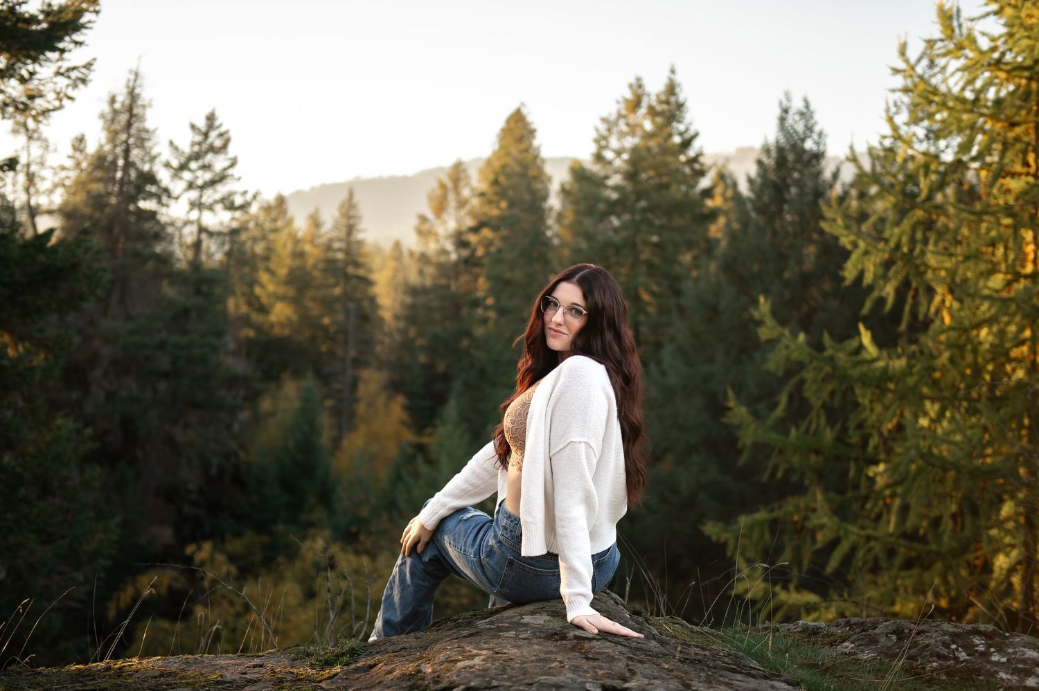 Young woman with long dark hair in white cardigan and jeans sitting on a rock surrounded by evergreen forest in Post Falls, ID.