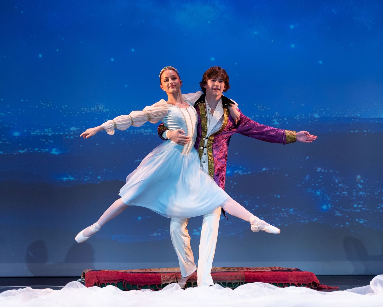 Two ballet dancers pose gracefully on a magic carpet set against a blue starry night stage backdrop.