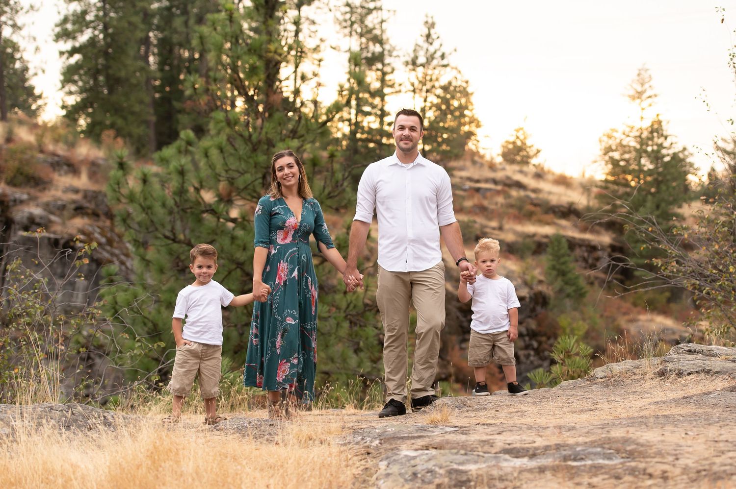 Smiling family of four walks hand-in-hand on a rocky trail surrounded by pine trees at sunset.