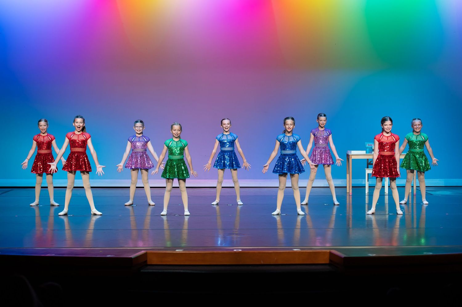 Young dance troupe performs on stage in red, purple, and green costumes under colorful lights.